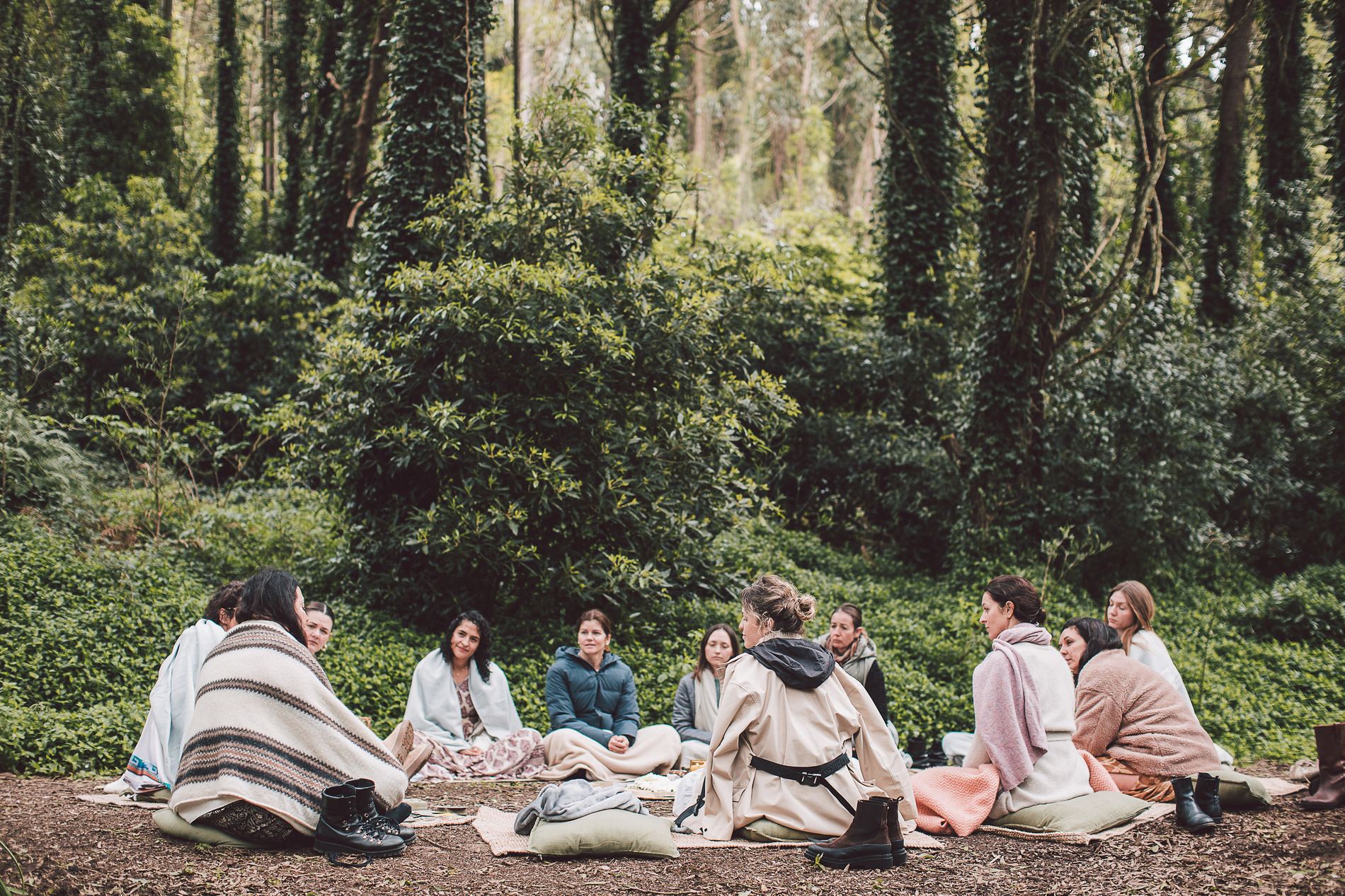 A circle of women sitting together on cushions in the forest, wrapped in blankets