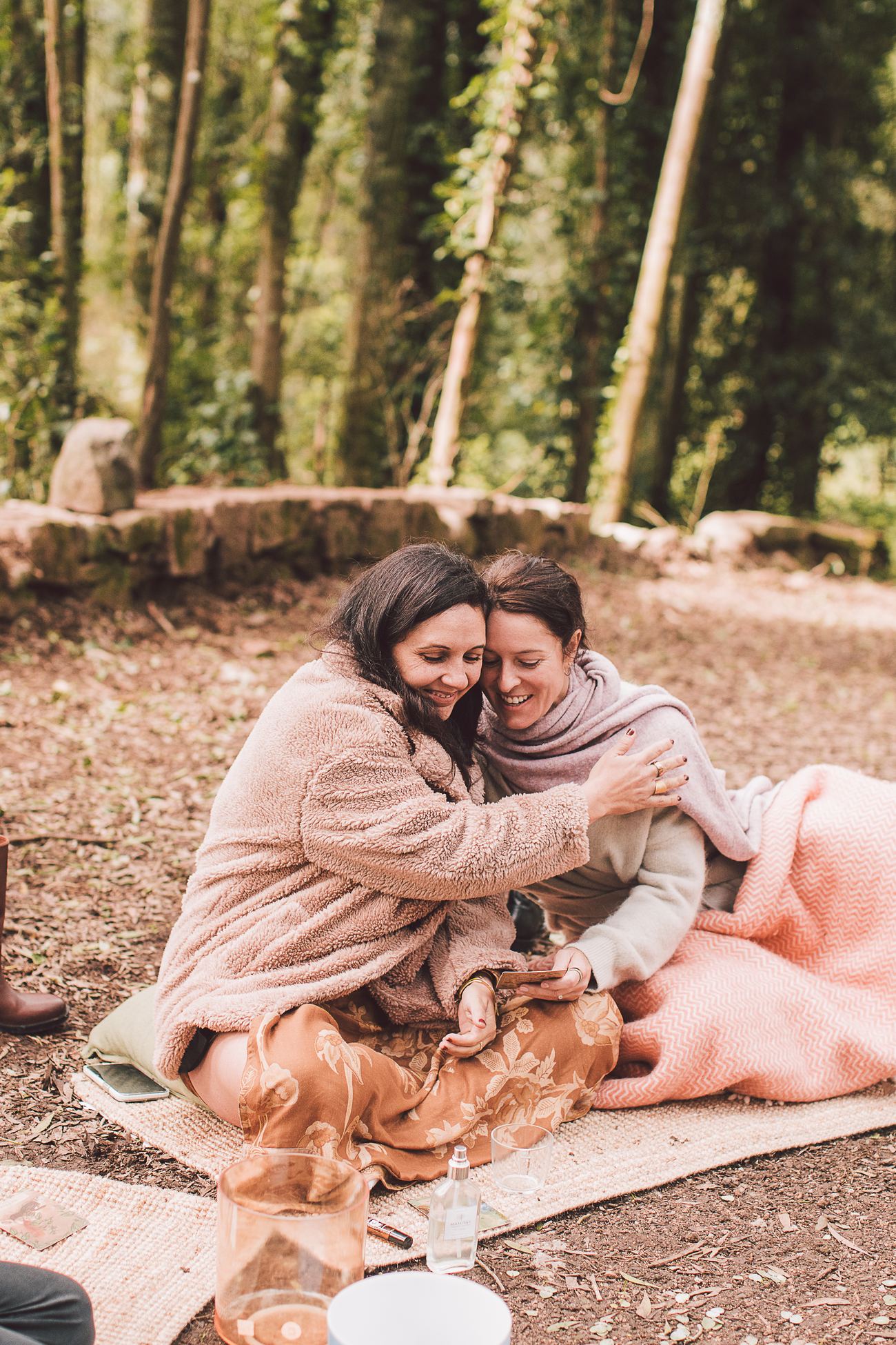 Two women sitting close together on the forest floor, laughing